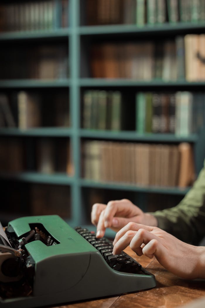 Close-up of hands typing on a vintage green typewriter in a library setting.