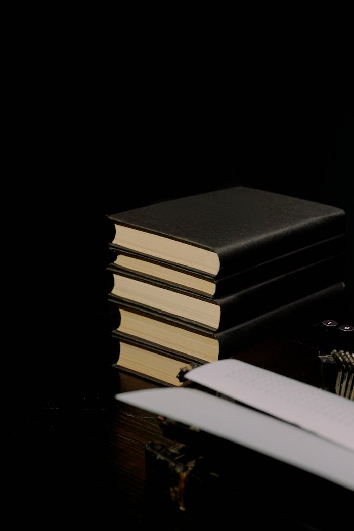 A stack of black hardcover books on a dark wooden table with a typewriter.